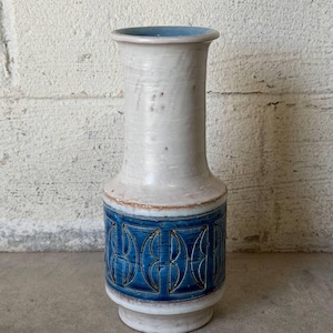 May include: A ceramic vase with a tall, slender neck and a wider base. The upper portion is off-white with subtle texture, while the lower section features a vibrant blue band decorated with a repeating geometric pattern of incised arches.