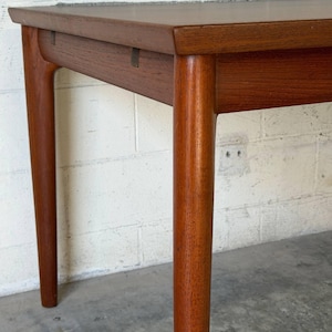 May include: A rectangular wooden table with a smooth, medium-brown finish. The table features four cylindrical, slightly tapered legs that connect to the apron with visible joinery details. The surface is flat with a clean, straight edge profile.