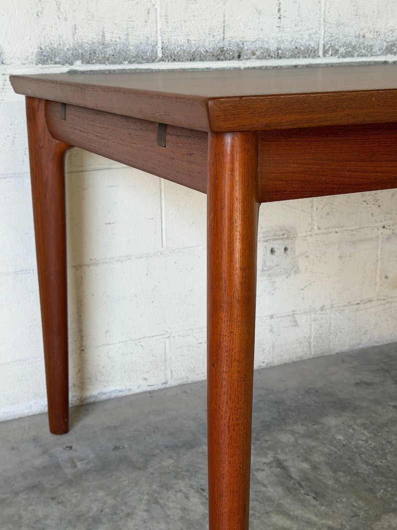 May include: A rectangular wooden table with a smooth, medium-brown finish. The table features four cylindrical, slightly tapered legs that connect to the apron with visible joinery details. The surface is flat with a clean, straight edge profile.