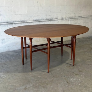 May include: An oval wooden dining table with a smooth, medium-brown finish. The table features a drop-leaf design supported by six slender, tapered legs connected by a structural cross-brace frame underneath the tabletop surface.