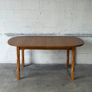 May include: A medium-brown wooden dining table with an oval top and four legs featuring a carved, segmented design. The table surface shows a visible wood grain pattern, and the structure is supported by four vertical legs with distinct decorative notches.