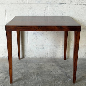 May include: A square wooden table with a dark brown finish and visible wood grain. The table features four straight, slightly tapered legs and a smooth, flat top surface with a subtle, natural wood knot detail along the front apron edge.