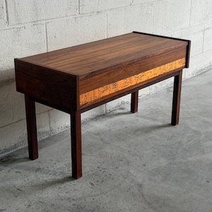 May include: A rectangular wooden console table with a dark brown wood grain finish. The front panel features a horizontal band of bright orange material decorated with black line drawings of abstract figures and symbols, supported by four legs.