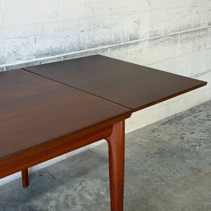 May include: A rectangular wooden dining table with a dark brown top and a lighter brown wooden frame. The table features a visible seam where an extension leaf meets the main surface, supported by four tapered legs extending to the floor.