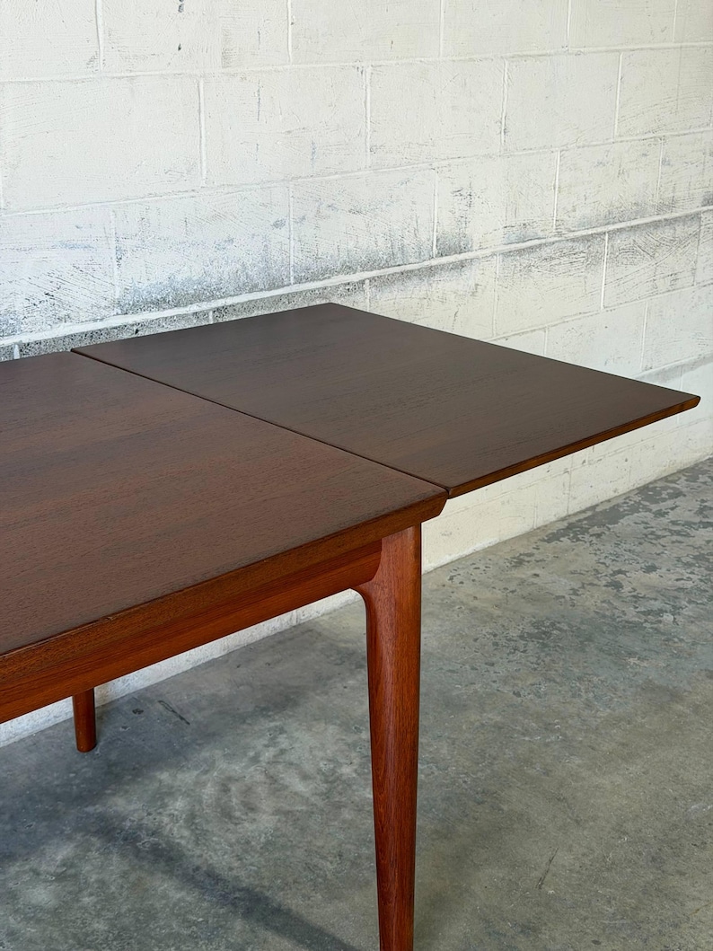 May include: A rectangular wooden dining table with a dark brown top and a lighter brown wooden frame. The table features a visible seam where an extension leaf meets the main surface, supported by four tapered legs extending to the floor.