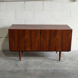 May include: A rectangular wooden sideboard featuring a rich, dark brown wood grain pattern with two cabinet doors that meet in the center. The furniture piece is supported by four tapered, cylindrical wooden legs positioned at each corner.