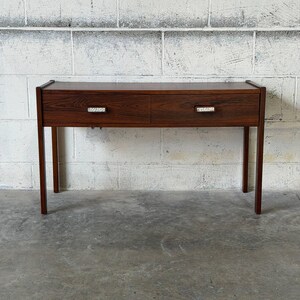 May include: A rectangular wooden console table featuring a dark brown wood grain finish. The table has four straight legs and two horizontal drawers positioned side by side, each equipped with a small, rectangular, textured metallic handle.
