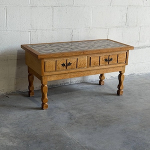May include: A rectangular wooden coffee table with a tiled top and three drawers. The table has four ornate legs and black drawer pulls. The top is a mosaic of gray and white tiles. The wood has a light brown finish.
