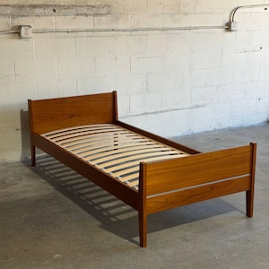 May include: A single bed frame constructed from medium-toned wood with a visible grain pattern. The frame features a tall headboard and a shorter footboard, both with straight lines, supported by four legs and a series of light-colored wooden slats.
