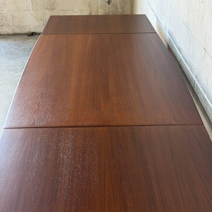 May include: A rectangular wooden dining table with a rich, dark brown finish and a visible wood grain pattern. The tabletop features a distinct seam across the centre, indicating an extendable design, supported by sturdy legs at the corners.