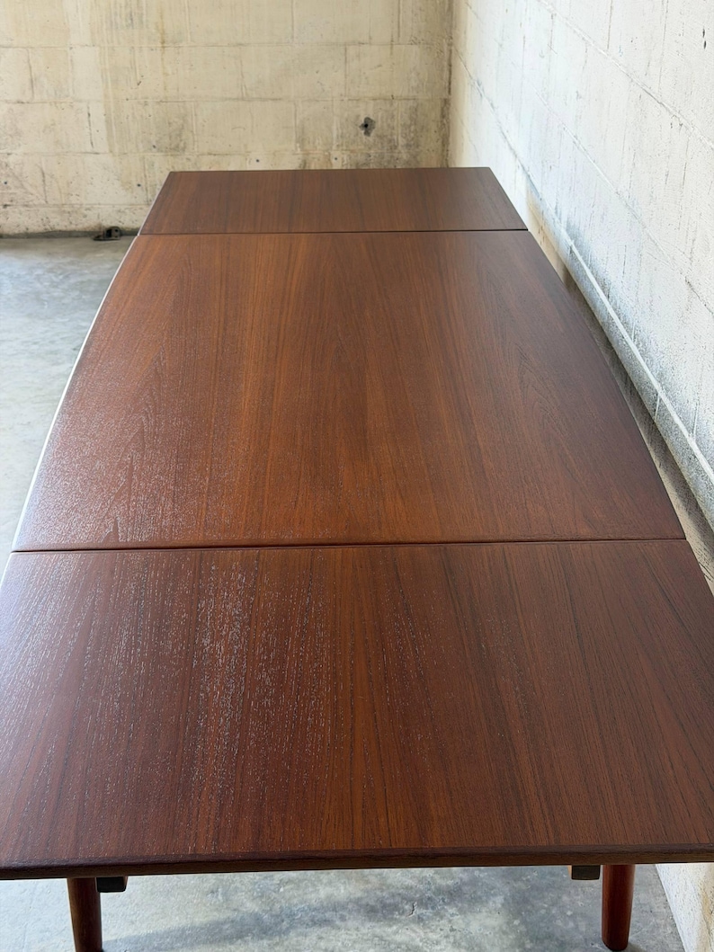 May include: A rectangular wooden dining table with a rich, dark brown finish and a visible wood grain pattern. The tabletop features a distinct seam across the centre, indicating an extendable design, supported by sturdy legs at the corners.