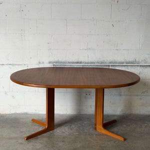 May include: An oval dining table made of warm-toned wood with a smooth, polished surface. The table features two vertical support columns, each connected to a pair of angled, splayed feet that extend outward to provide a stable base.