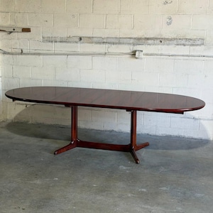 May include: A large, oval-shaped dining table with a deep reddish-brown wood grain finish. The table features a smooth, polished surface and is supported by a central pedestal base with two splayed feet extending outward on each side.