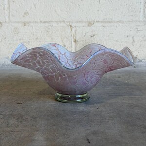 May include: A decorative glass bowl with a ruffled rim. The bowl is a soft pink color with a textured pattern and a clear green base. The rim has a light blue edge.