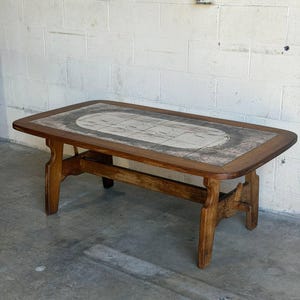 May include: A rectangular coffee table with a medium-toned wooden frame and four sturdy legs. The tabletop features a central inlay of rectangular ceramic tiles arranged in a grid pattern, displaying a muted gray and white abstract design.
