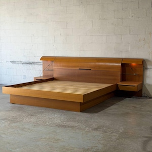 May include: A wooden bed frame featuring a wide, curved headboard with integrated side tables and glass shelving. The bed includes a low-profile platform base with wooden slats and built-in lighting fixtures on both sides of the headboard.