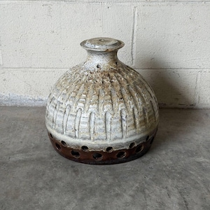 May include: A round ceramic vessel with a narrow neck and a flat rim. The upper portion features a textured, cream-colored glaze with vertical indentations, while the lower base is a smooth, dark brown band punctuated by a series of circular holes.