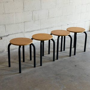 May include: Four wooden stools with round, light-brown seats and black metal legs. The legs have a curved design. The stools are arranged in a row against a white brick wall and a gray concrete floor.
