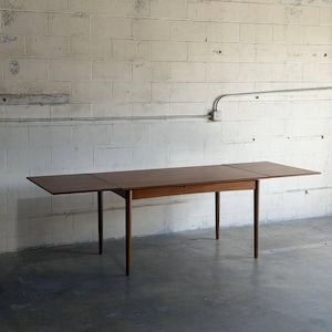 May include: A rectangular wooden dining table with a warm brown finish and a smooth surface. The table features four tapered cylindrical legs and two extendable leaves on either side, creating a long, flat surface for dining or workspace needs.