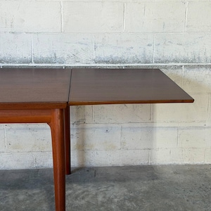 May include: A rectangular wooden dining table with a smooth, dark brown surface and a visible seam indicating an extendable leaf. The table features sturdy, tapered legs made of a lighter-toned wood, providing a stable base for the tabletop.