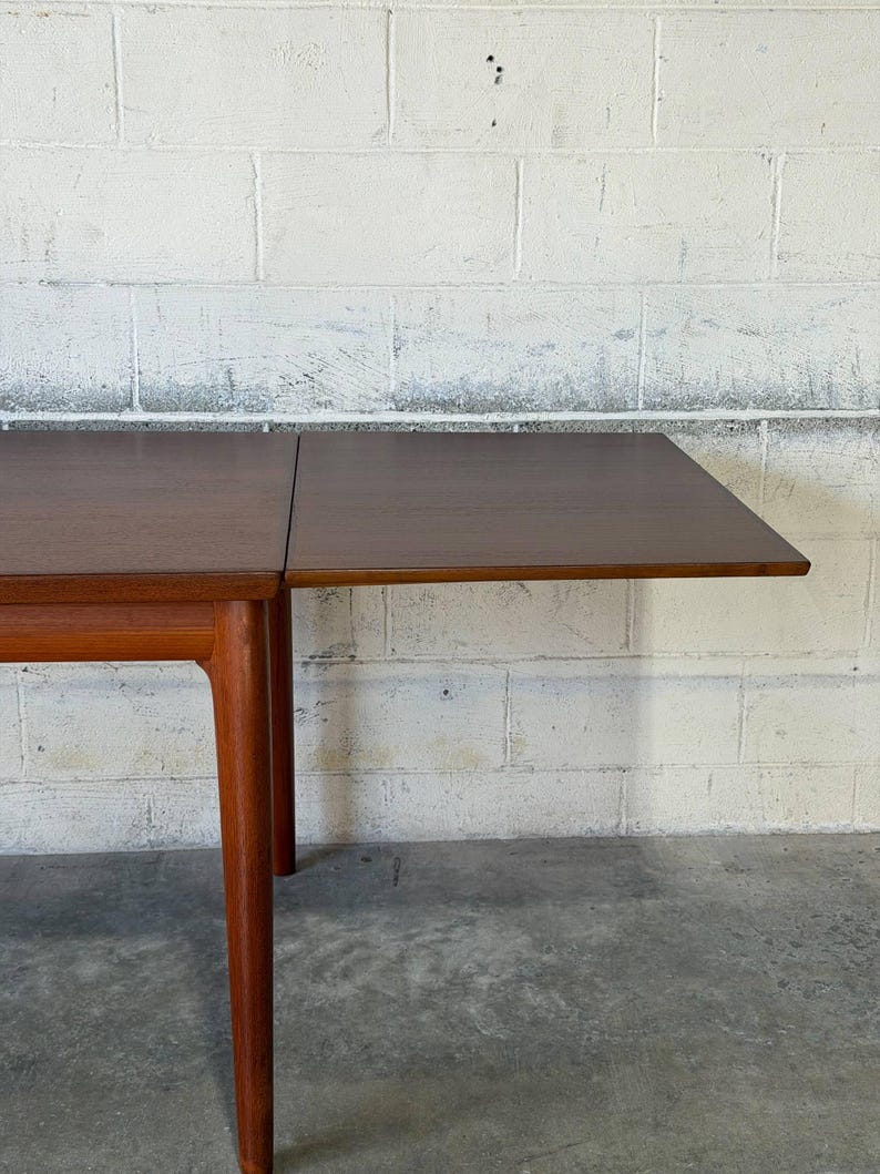 May include: A rectangular wooden dining table with a smooth, dark brown surface and a visible seam indicating an extendable leaf. The table features sturdy, tapered legs made of a lighter-toned wood, providing a stable base for the tabletop.
