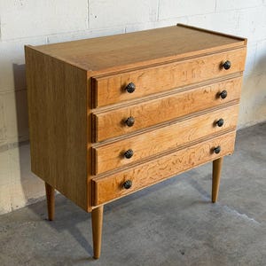 May include: A light brown wooden dresser featuring four horizontal drawers with a visible wood grain pattern. Each drawer is equipped with two small, dark, round, textured knobs. The piece is supported by four tapered wooden legs.