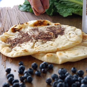 May include: Two flatbreads topped with zaatar seasoning, surrounded by blueberries, uncooked noodles, and a bag of zaatar seasoning. The image shows a hand sprinkling seasoning on the flatbreads. The food items are arranged on a wooden surface.