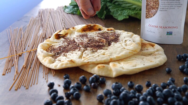 May include: Two flatbreads topped with zaatar seasoning, surrounded by blueberries, uncooked noodles, and a bag of zaatar seasoning. The image shows a hand sprinkling seasoning on the flatbreads. The food items are arranged on a wooden surface.