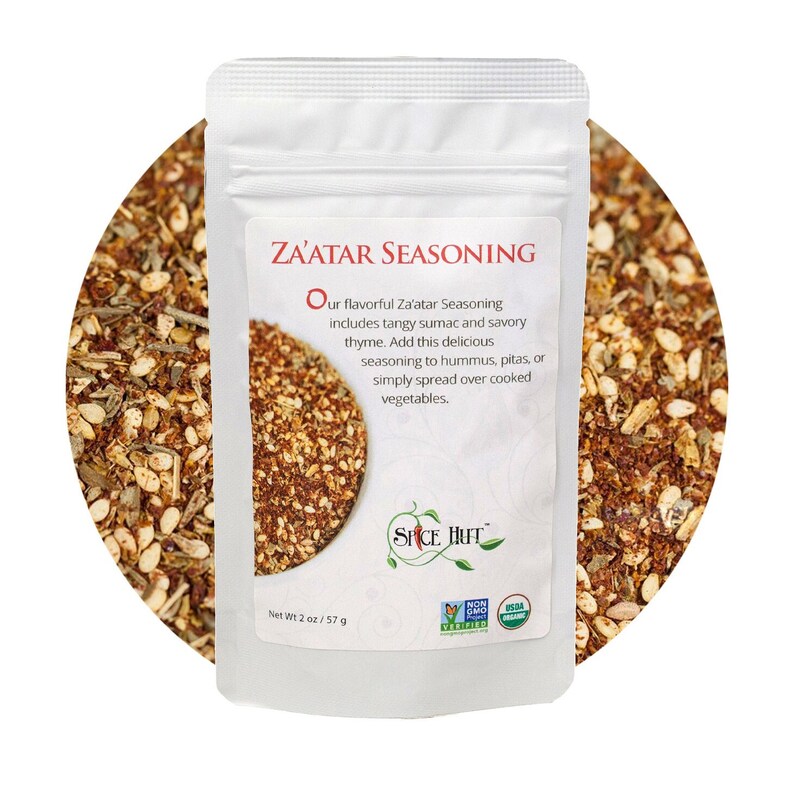 May include: A white pouch of Za'atar seasoning with text. The label reads "Za'atar Seasoning" and describes the ingredients and uses. The pouch is set against a background of the seasoning mix. Net weight is 2 oz / 57 g.