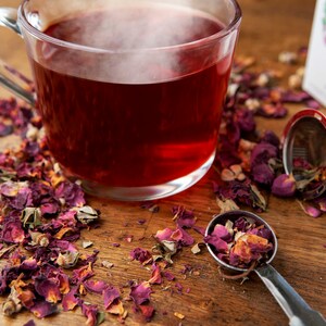May include: A glass cup filled with red rose tea, with loose rose petals scattered around the cup and a metal tea infuser on a wooden surface.