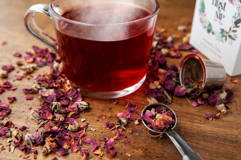 May include: A glass cup filled with red rose tea, with loose rose petals scattered around the cup and a metal tea infuser on a wooden surface.