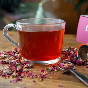May include: A glass mug filled with red rose tea, surrounded by loose rose petals and a tea infuser. The tea bag box is in the background with the text "The First Sip of Tea".
