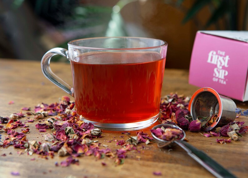 May include: A glass mug filled with red rose tea, surrounded by loose rose petals and a tea infuser. The tea bag box is in the background with the text "The First Sip of Tea".
