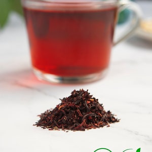 May include: A pile of dried hibiscus flowers, a cup of hibiscus tea, and two labels. One label says "Vegan" and the other says "100% natural ingredients".