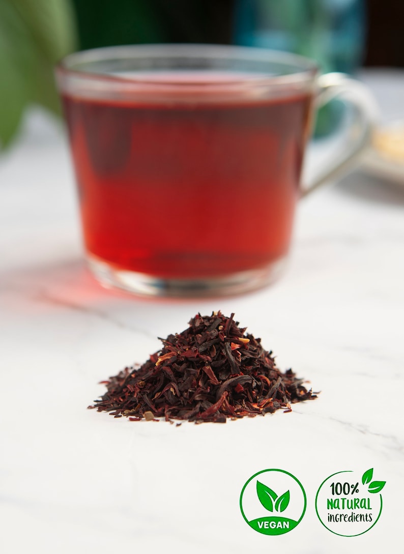 May include: A pile of dried hibiscus flowers, a cup of hibiscus tea, and two labels. One label says "Vegan" and the other says "100% natural ingredients".