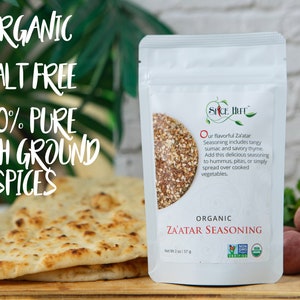 May include: A white pouch of organic Za'atar seasoning with text that reads "Organic Salt Free 100% Pure Fresh Ground Spices". The pouch is next to a stack of flatbread and a selection of fresh vegetables.