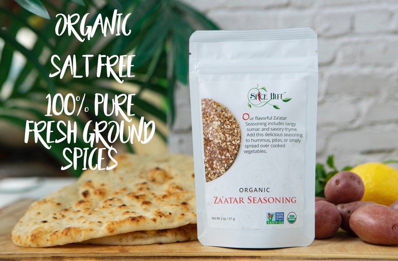 May include: A white pouch of organic Za'atar seasoning with text that reads "Organic Salt Free 100% Pure Fresh Ground Spices". The pouch is next to a stack of flatbread and a selection of fresh vegetables.