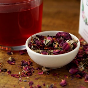 May include: A white bowl filled with loose leaf tea, including dried rose petals, green tea leaves, and other herbs. The tea is scattered on a wooden surface next to a glass mug of red tea and a box of tea labeled "The First Sip".