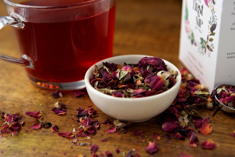 May include: A white bowl filled with loose leaf tea, including dried rose petals, green tea leaves, and other herbs. The tea is scattered on a wooden surface next to a glass mug of red tea and a box of tea labeled "The First Sip".