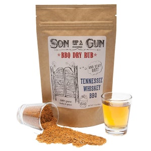 Puede incluir: Una bolsa de papel marrón con una etiqueta blanca que dice "Son of a Gun Seasonings *BBQ Dry Rub*" con un dibujo de una puerta de saloon y el texto "Tennessee Whiskey BBQ" y "100% pure herbs & spices". Hay un pequeño vaso de licor ámbar y una taza medidora de vidrio con un frotamiento de especias marrón.