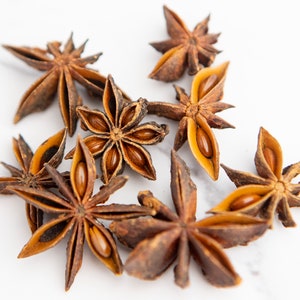 May include: A close-up of eight brown star anise pods on a white surface. The pods are open and show the seeds inside.