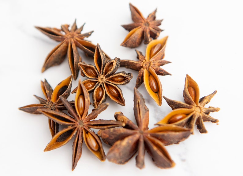 May include: A close-up of eight brown star anise pods on a white surface. The pods are open and show the seeds inside.