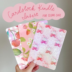 May include: Three pink and white patterned cardstock inserts for a clear Kindle case. The inserts feature a repeating pattern of cherries and strawberries. The text "Cardstock Kindle Insert for Clear Case" is written in pink on a white cloud shape.