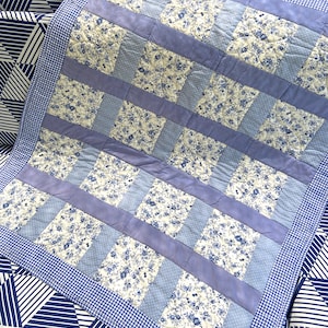 May include: A blue and white patchwork quilt with a floral pattern. The quilt is made of squares of different fabrics, some with a floral print and some with a solid colour. The quilt is edged with a blue and white gingham border.