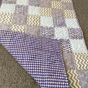 May include: A baby quilt with a patchwork design featuring various geometric patterns in shades of grey, blue, and yellow. The back of the quilt is a navy blue and white houndstooth pattern.