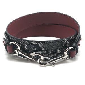 May include: A black and white snakeskin patterned leather strap with a burgundy leather backing. The strap has silver metal hardware with two hooks.