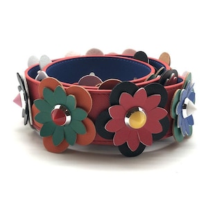May include: A red leather bracelet with colorful flower appliqués. The flowers are in shades of green, orange, black, red, and blue, each with a silver stud center. The bracelet has a blue interior and is set against a white background.