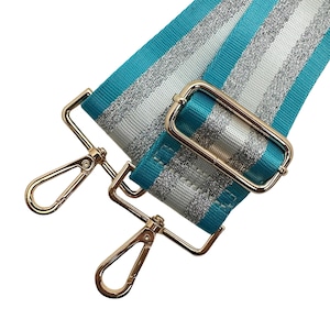 Peut inclure: Une bandouli&egrave;re de sac &agrave; rayures turquoise, argent et blanches avec une quincaillerie en m&eacute;tal dor&eacute;. La sangle pr&eacute;sente un motif de rayures r&eacute;p&eacute;t&eacute;es et comprend deux mousquetons pivotants et une boucle rectangulaire.