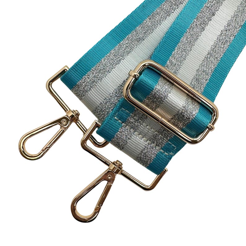 Peut inclure: Une bandouli&egrave;re de sac &agrave; rayures turquoise, argent et blanches avec une quincaillerie en m&eacute;tal dor&eacute;. La sangle pr&eacute;sente un motif de rayures r&eacute;p&eacute;t&eacute;es et comprend deux mousquetons pivotants et une boucle rectangulaire.