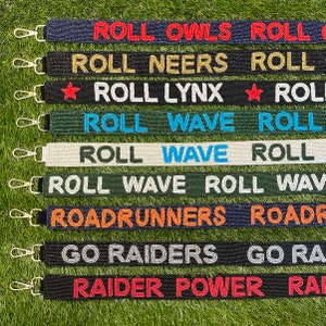 May include: A set of nine beaded keychains with different colors and text. The keychains feature the words "Roll Owls Roll", "Roll Neers Roll", "Roll Lynx", "Roll Wave Roll", "Roadrunners", "Go Raiders", and "Raider Power".